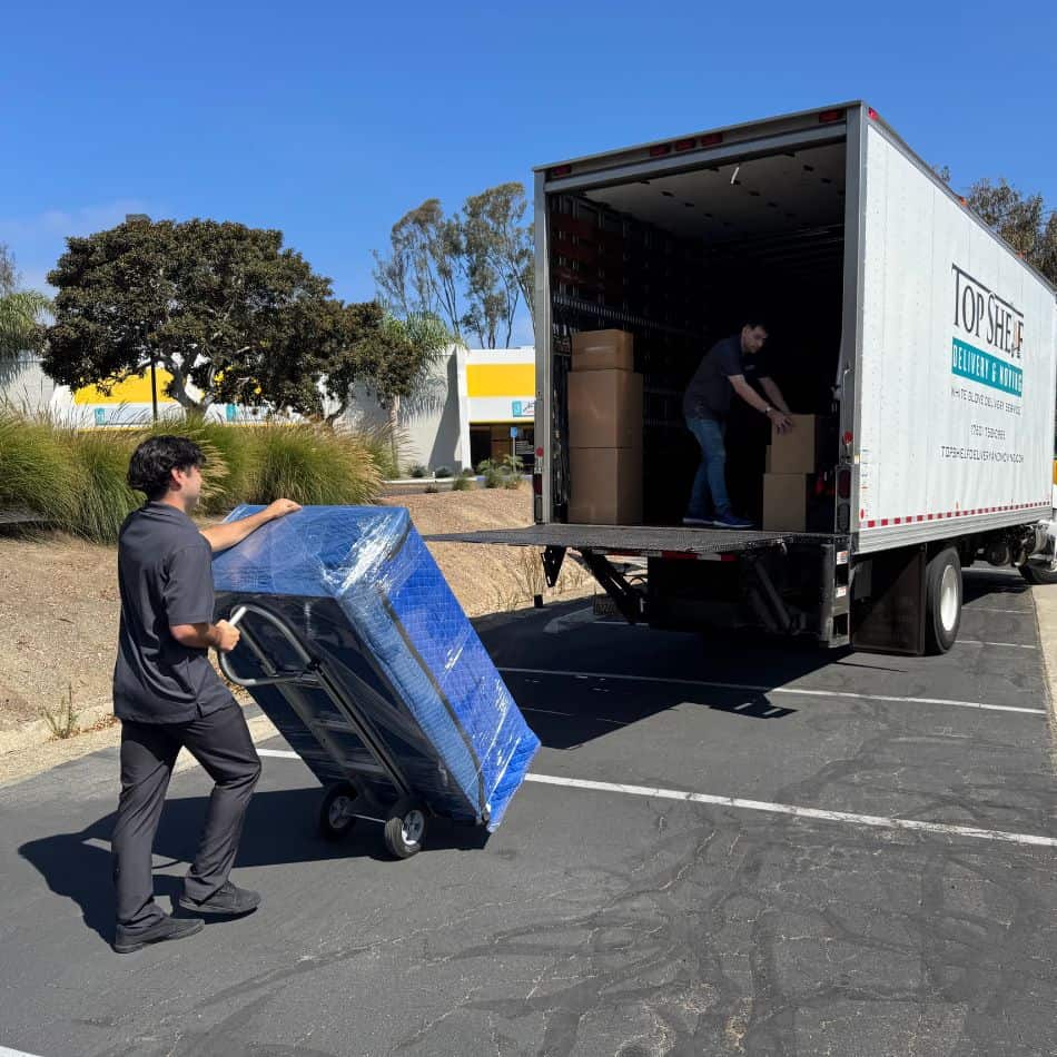 Professional mover using a heavy-duty dolly to load a blue-wrapped furniture item into a Top Shelf moving truck.
