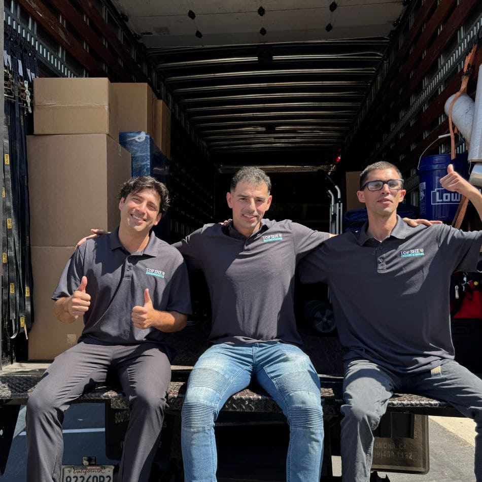 Three smiling Top Shelf movers sitting on the liftgate of a fully organized moving truck packed with professional crates.